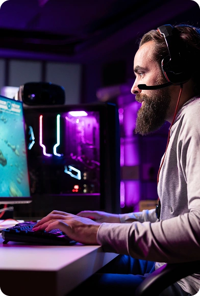 Een man met een baard en een headset speelt intensief een videogame op een breedbeeldscherm, in een schemerige kamer met neonverlichting en een hoogwaardige gaming-opstelling